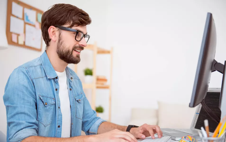 Java developer with a beard, wearing a blue shirt and a white T-shirt working on a computer